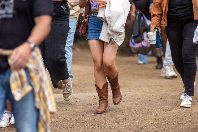 People walk on a dirt path, with one person wearing brown cowboy boots and a blue skirt holding a jacket, while others wear casual shoes and carry items.